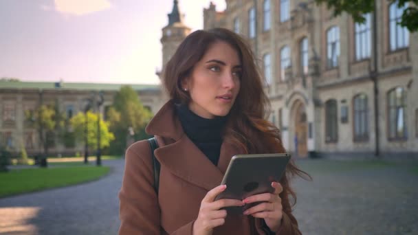 Carino caucasico ragazza bruna è in piedi rilassato e tenendo tablet con le mani, guardando e utilizzando il suo dispositivo, illustrazione urbana, persona moderna in sole — Video Stock