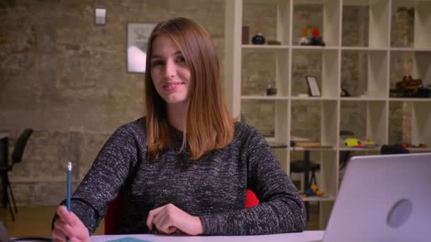 Redheaded young woman smilingly watches into camera and interacts with pensil in the office — Stock Video