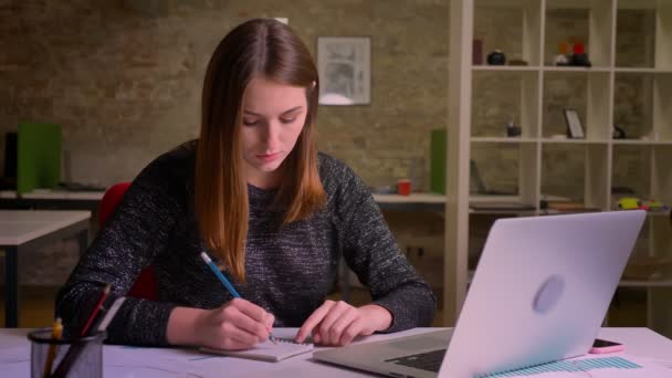 युवा लाल बालों वाली महिला लैपटॉप के सामने काम कर रही है और कार्यालय में पेंसिल और नोटबुक का उपयोग करके नोट्स बना रही है — स्टॉक वीडियो