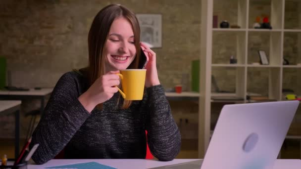 Chica joven pelirroja en el lugar de trabajo habla alegremente a través del teléfono móvil y bebidas . — Vídeos de Stock