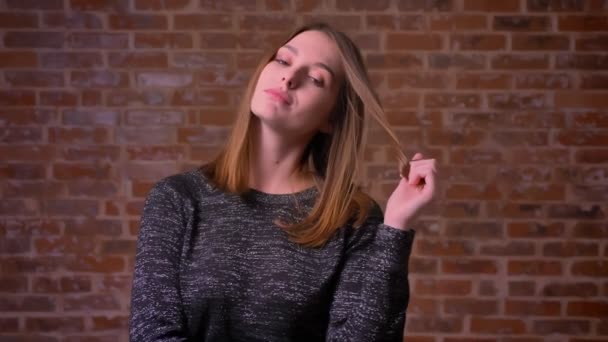 Ginger young woman flirting with the camera and twisting her hair on the finger on a bricken background. — Stock Video