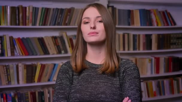 Kaukázusi vörös hajú diáklány néz a kamerába komolyan és a flörtölni a könyvespolcok háttér. — Stock videók