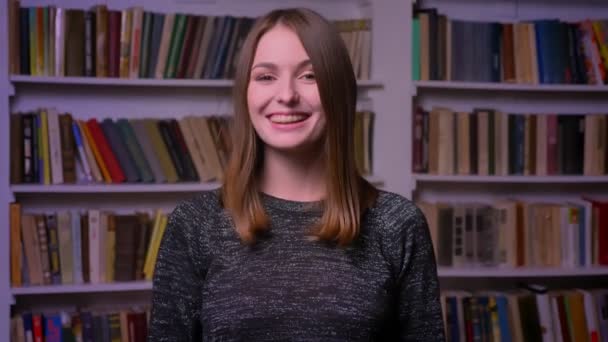 Roodharige Kaukasische meisje in bretels horloges glimlachend in de camera, vinger haar tot Toon zoals en respect op boekenkasten achtergrond — Stockvideo