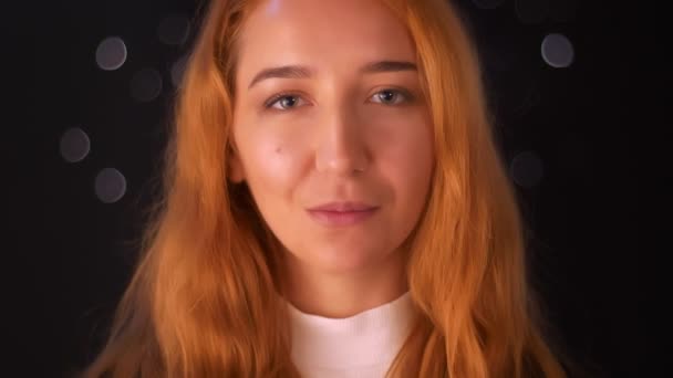 Восхитительные кадры крупным планом кавказская рыжая женщина стоит и смотрит в камеру с естественной улыбкой и открытым взглядом, спокойно, темная студия на заднем плане, прекрасная поза — стоковое видео