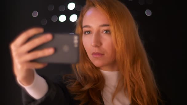 Underbara kaukasisk kvinna tar selfie på sin telefon medan stående isolerad på mörk bakgrund med whil espont på det, tittar på skärmen avslappnad och njuter av chill tid — Stockvideo