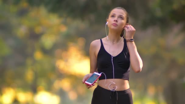 Close-up retrato de menina em macacão colocando fones de ouvido e começando a correr no parque outonal — Vídeo de Stock