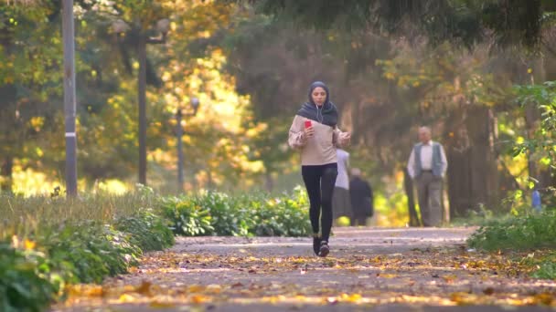 Muslimsk kvinna i hijab med hörlurar går jogging i höstlig park med människor på bakgrunden — Stockvideo