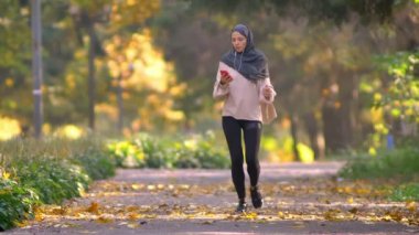 Hijab Müslüman kadın sonbahar parkta koşu gider, smartphone bakmak için durdurur ve çalışmaya devam eder