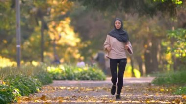 Hijab Müslüman kadında sonbahar parkta koşu gider, saati kontrol etmek için durdurur ve çalışmaya devam eder