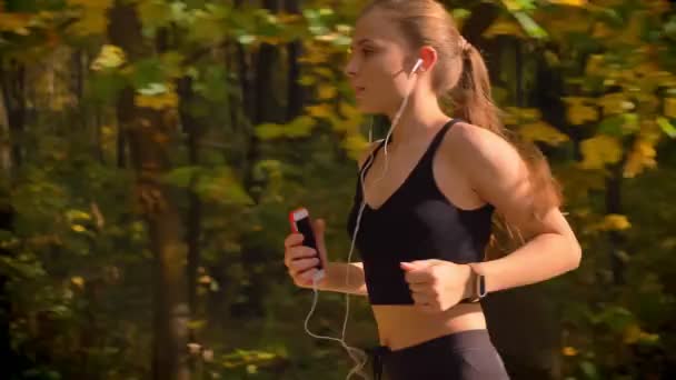 在秋天的公园里带着电话和耳机奔跑的年轻白种人苗条女孩的多利镜头 — 图库视频影像