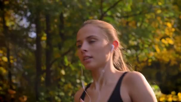 ドリー ショット、イヤホンで秋の公園を実行する若い魅力的な白人スリムな女の子のクローズ アップの肖像画. — ストック動画