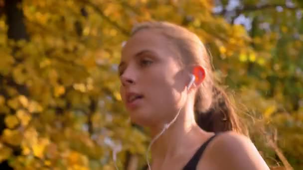 Dolly shot from face to chest, close-up portrait of young caucasian slim girl running in autumnal park with earphones. — Stock Video