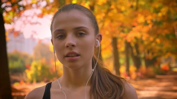 Gros plan portrait de jeune fille caucasienne avec écouteurs regarder dans la caméra en essayant de reprendre son souffle après avoir couru . — Video