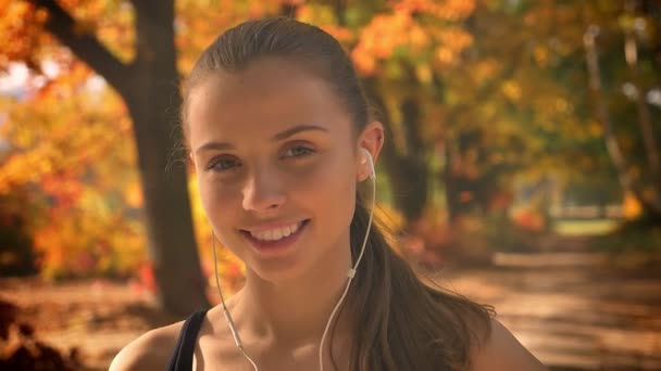 Giovane ragazza caucasica con gli auricolari si rivolge alla fotocamera e sorride su sfondo autunnale . — Video Stock