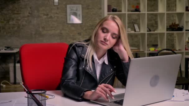 Mooie Kaukasische blonde vrouw is zittend op bureaublad en ontspannen, met werk breken in de buurt van haar laptop en documenten, licht baksteen office op achtergrond — Stockvideo