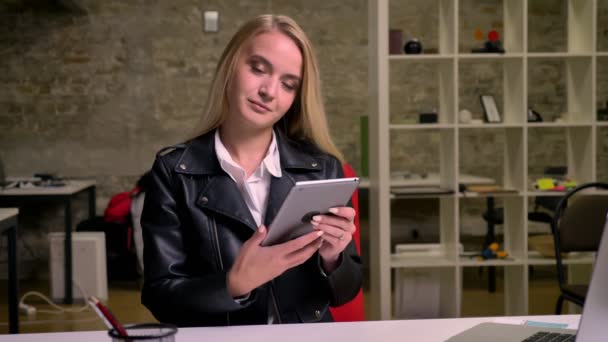 Гарненька блондинка біла жінка в офісі сидить і показує зелений екран на своєму планшеті з приємною посмішкою на обличчі і виглядає холодною на камеру, офісний фон — стокове відео