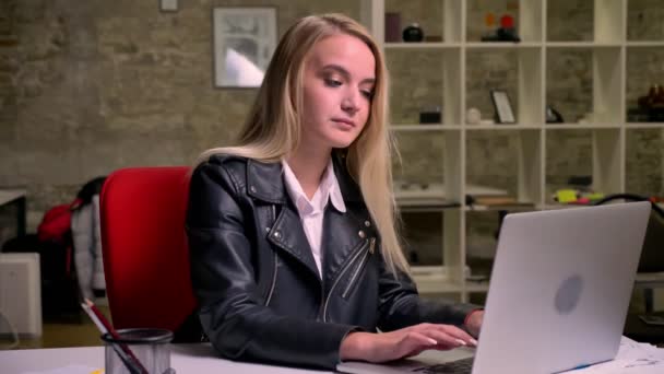 Досить біла блондинка зв'язується на клавіатурі ноутбука і рухається розслабленим прослуховуванням музики, сидячи на робочому столі в зеленому цегляному офісі — стокове відео