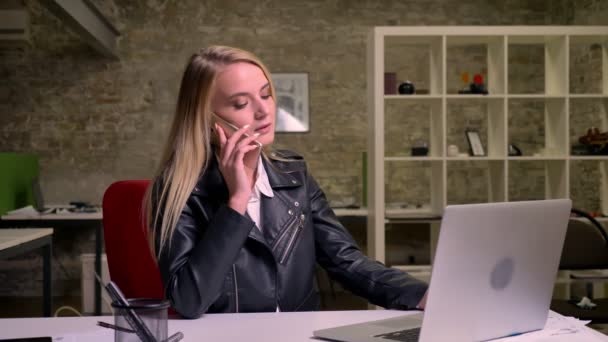 Красива блондинка біла бізнес-леді висить по телефону зі спокійним концентрованим обличчям, сидячи на робочому столі біля ноутбука в цегляному офісі — стокове відео