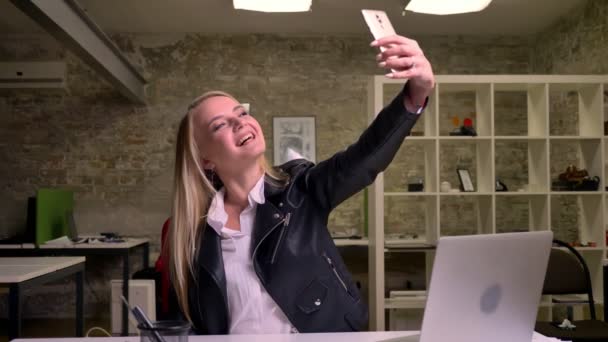 Glücklich lächelnde kaukasische Blondine sitzt am Schreibtisch und macht Selfies mit ihrem Smartphone, während sie im hellen Backsteinbüro hängt — Stockvideo