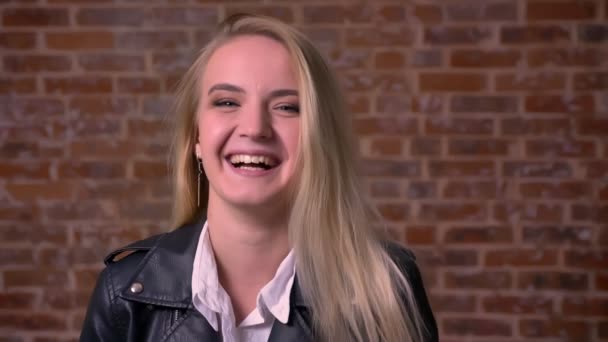 Artistic blonde caucasian female close-up standing and laughing at camera, relaxed and happy, isolate don brick red wall, indoors — Stock Video