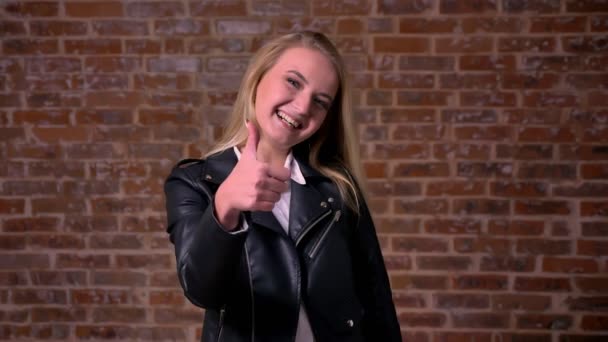 Pretty blonde caucasian woman in black jacket is standing straight and showing like sign at camera chill and smiling cheerfully, red brick wall behind — Stock Video