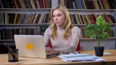 Belgili tanımlık laptop önünde çalışma ve kitap rafı arka plan üzerinde notlar yapma sarışın beyaz kadın.