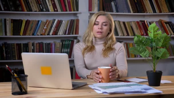 Porträt einer blonden Frau mittleren Alters, die Kaffee trinkt und auf dem Bildschirm des Laptops im Bücherregal zusieht. — Stockvideo
