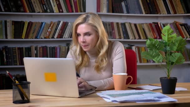Portrait de femme caucasienne blonde d'âge moyen travaillant avec un ordinateur portable et réalisant un succès sur fond de bibliothèques . — Video