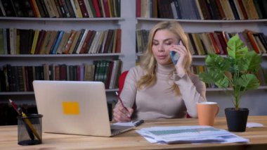 Telefonda konuşmak ve Office kitap rafı arka plan üzerinde notlar yapma orta yaşlı beyaz sarışın portresi.