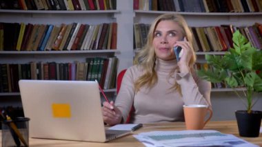 Smartphone üzerinde konuşmak ve ofis onun koltukta kitap rafı arka plan üzerinde değişen sarışın beyaz iş kadını.