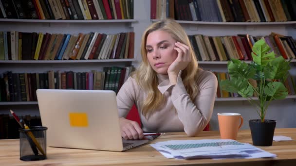 Porträtt av uttråkade blonda medelålders kaukasiska kvinna att skriva med laptop i office på bokhyllor bakgrund. — Stockvideo