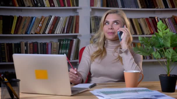 Rubia mujer de negocios caucásica hablando en el teléfono inteligente y cambiando en su asiento en la oficina en los estantes de fondo . — Vídeo de stock