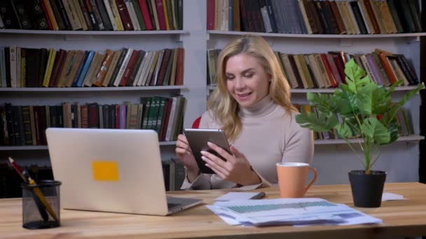 Blondine mittleren Alters kaukasische Geschäftsfrau arbeitet mit Tablet im Büro auf Bücherregalen Hintergrund. — Stockvideo