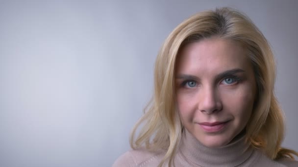 Close-up portrait of middle-aged blonde lady raising her eyes and smiling into camera on gray background. — Stock Video