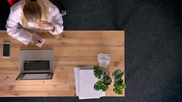 Dolly toma de vista de arriba hacia abajo de la mujer vestida formalmente trabajando y tomando notas en el cuaderno en la oficina delante de la computadora portátil . — Vídeo de stock