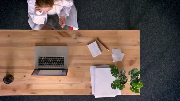 Dolly shot, vista dall'alto verso il basso di una donna rossiccia vestita con camice da laboratorio che parla al cellulare in ufficio davanti al portatile . — Video Stock