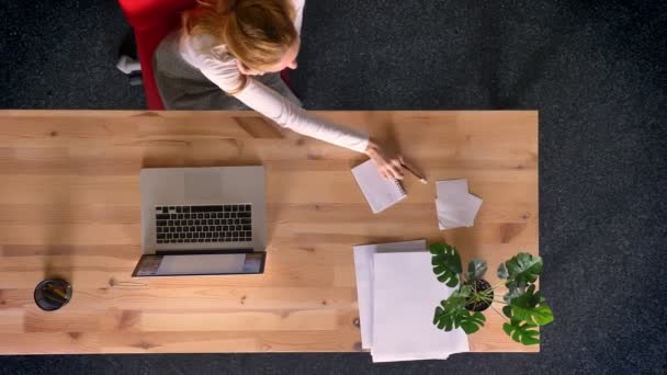 Dolly shot, top down view of redheaded woman working with computer, talking on cellphone and making notes — Stock Video