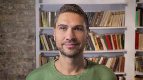 Homem adulto olhando para a frente, sorrindo e piscando calmamente. Estantes de livros em segundo plano — Vídeo de Stock