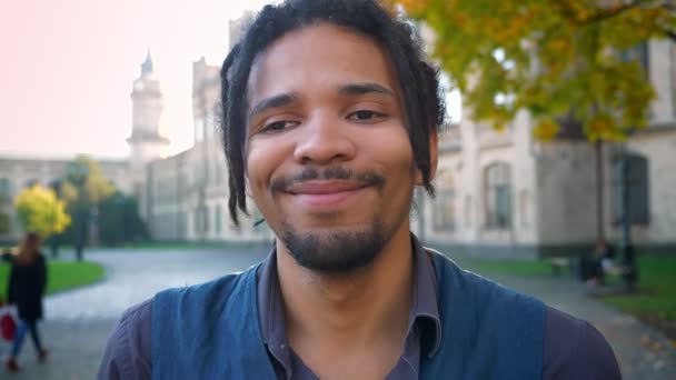 Крупный план портрета молодого афроамериканца с дредами, скромно смеющегося в камеру на университетском фоне . — стоковое видео