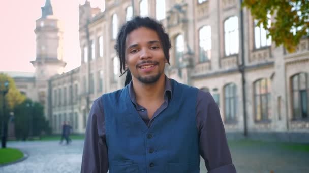 Portrait de jeune afro-américain avec dreadlocks riant humblement dans la caméra sur fond universitaire . — Video