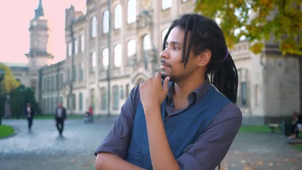 Close-up retrato de estudante afro-americano com dreadlocks tocando atentamente sua barba no fundo da universidade . — Vídeo de Stock