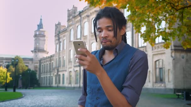 Retrato de estudiante afroamericano con rastas examinando emocionalmente el contenido en un teléfono inteligente con antecedentes universitarios . — Vídeos de Stock