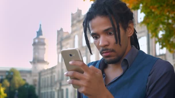 Portrait d'un étudiant afro-américain avec dreadlocks regardant du contenu au téléphone avec amusement sur fond universitaire . — Video