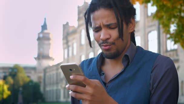 Πορτρέτο του Αφρικανικός-Αμερικανός φοιτητής με dreadlocks βλέποντας περιεχόμενο στο τηλέφωνο με ικανοποίηση στο Πανεπιστήμιο φόντο. — Αρχείο Βίντεο