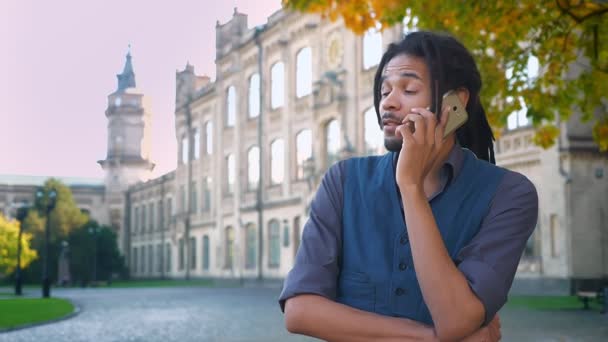 Portrait of attractive African-American student with dreadlocks emotionally talking on cellphone on university background. — Stock Video