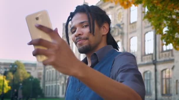 Close-up portrait of African-American student with dreadlocks posing to make selfies on smartphone on university background. — Stock Video