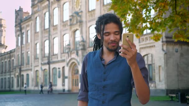 Estudante afro-americano atraente com dreadlocks falando em videochat usando smartphone em fundo universitário . — Vídeo de Stock