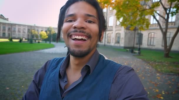 Selfie-vídeo de jovem estudante afro-americano com dreadlocks falando em videochat sobre fundo universitário . — Vídeo de Stock