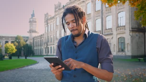 Retrato de un atractivo estudiante afroamericano con rastas trabajando con tableta sobre fondo universitario . — Vídeos de Stock