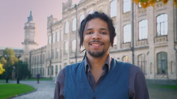 Retrato de atractivo afroamericano con rastas sonriendo a la cámara sobre fondo universitario . — Vídeos de Stock